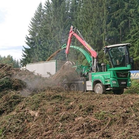 Eine grüne Maschine bearbeitet einen Holzstapel auf einem Baustellenareal.